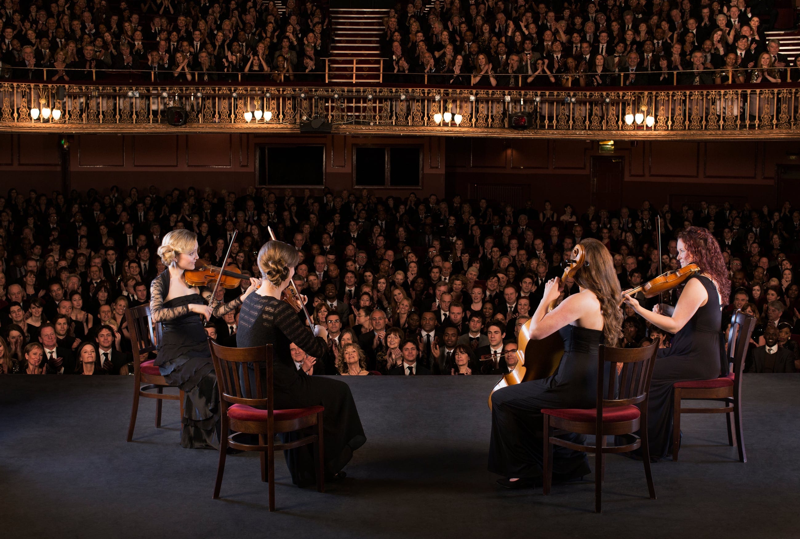 A string quartet on a theater stage before a full house — rows of listeners leaning in, faces carrying anticipation